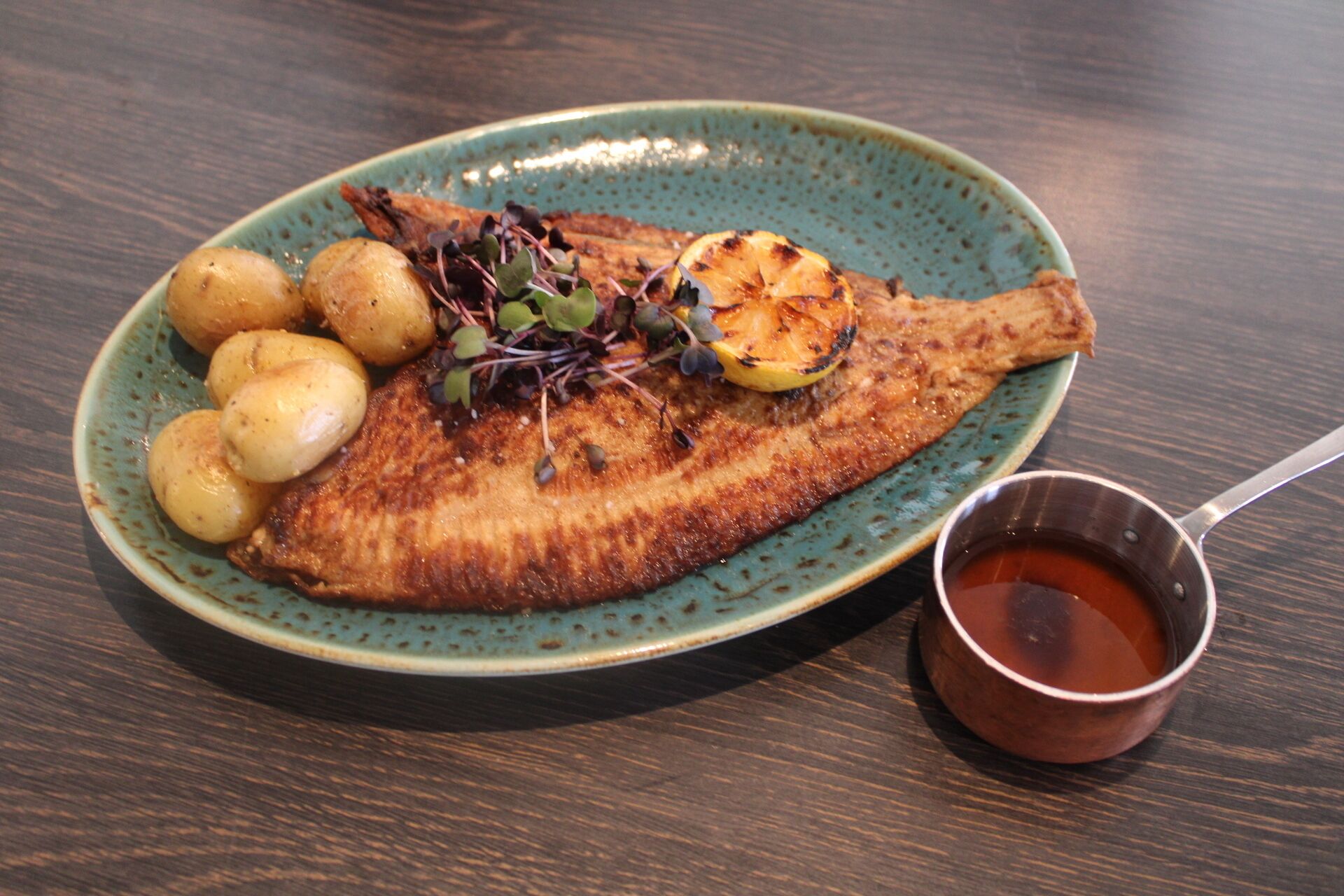 Un plato con pescado a la parrilla, patatas y una guarnición, acompañado de un recipiente pequeño con salsa.