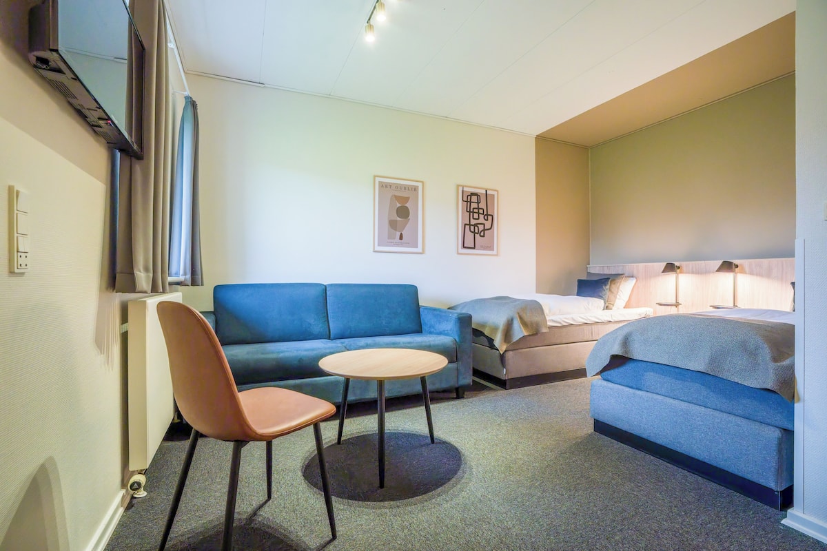 A hotel room with a blue sofa, a small round table, a chair, a bed, and two wall-mounted lamps.