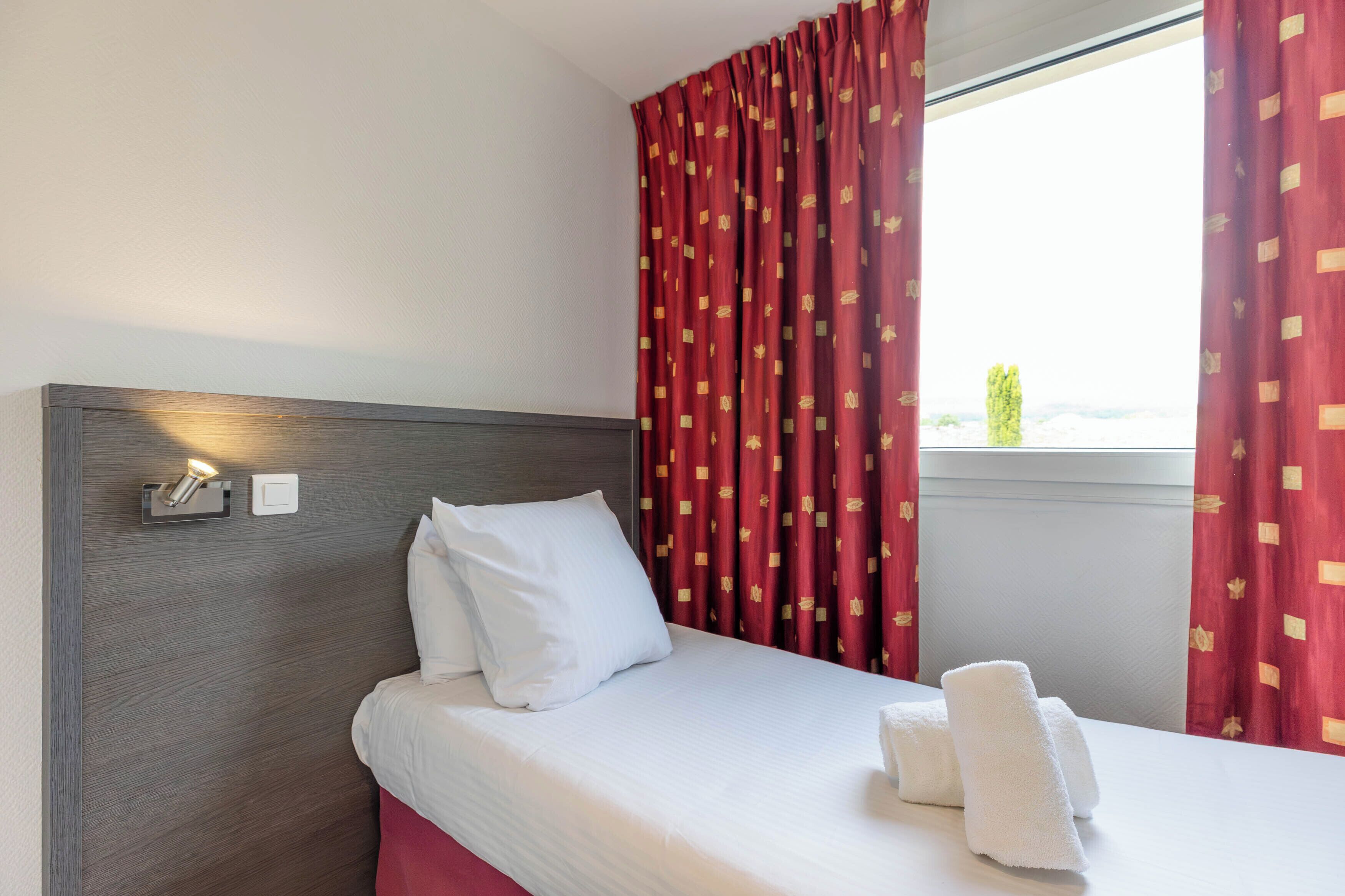A hotel room with a bed, white pillows, a headboard, a nightstand with a lamp, and red curtains.