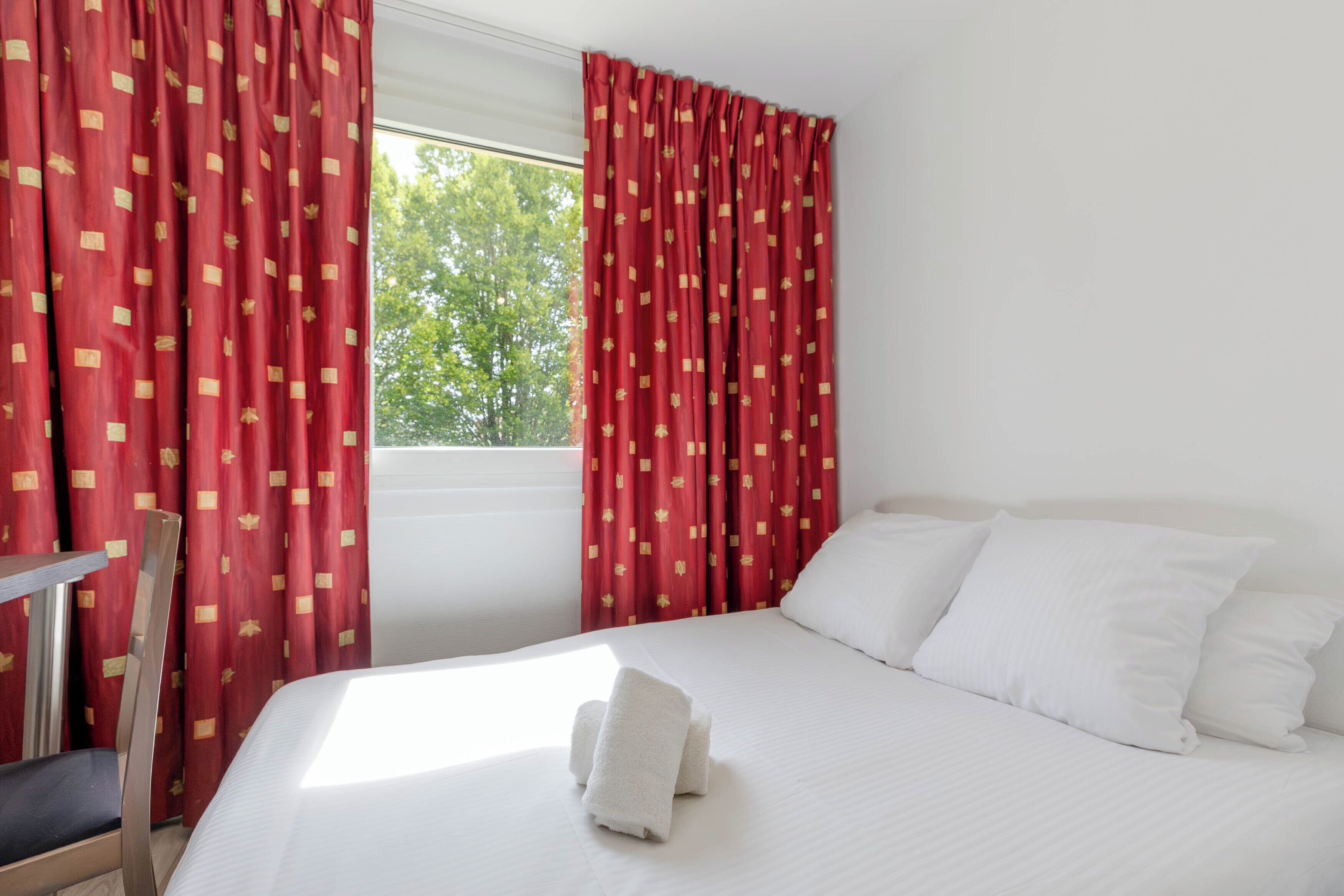 Habitación de hotel con una cama, almohadas blancas, cortinas rojas con estampado y una ventana con vista a árboles.