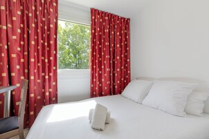 Habitación de hotel con una cama, almohadas blancas, cortinas rojas con estampado y una ventana con vista a árboles.