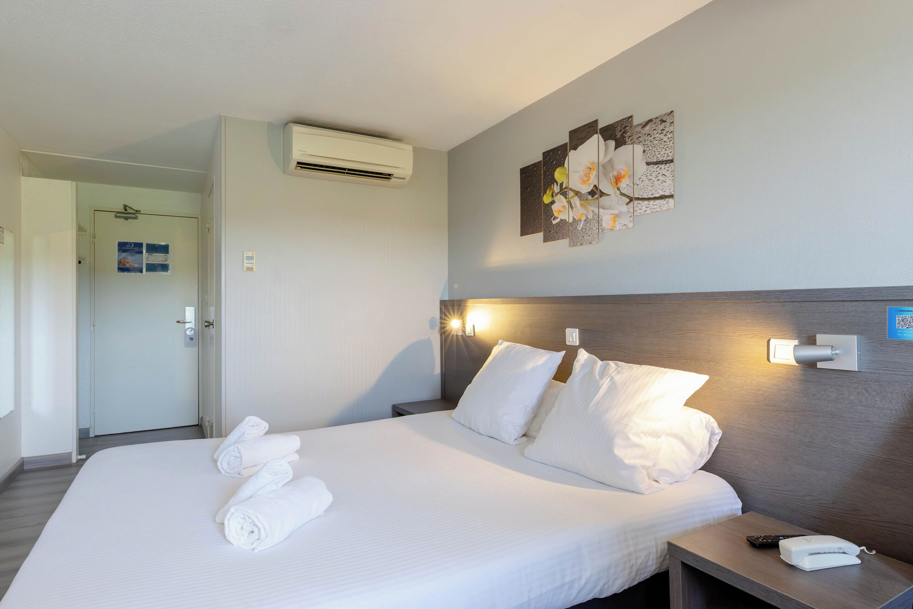 A hotel room with a bed, pillows, a nightstand with a phone, an air conditioner, and a wall-mounted light fixture.