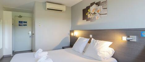 A hotel room with a bed, pillows, a nightstand with a phone, an air conditioner, and a wall-mounted light fixture.