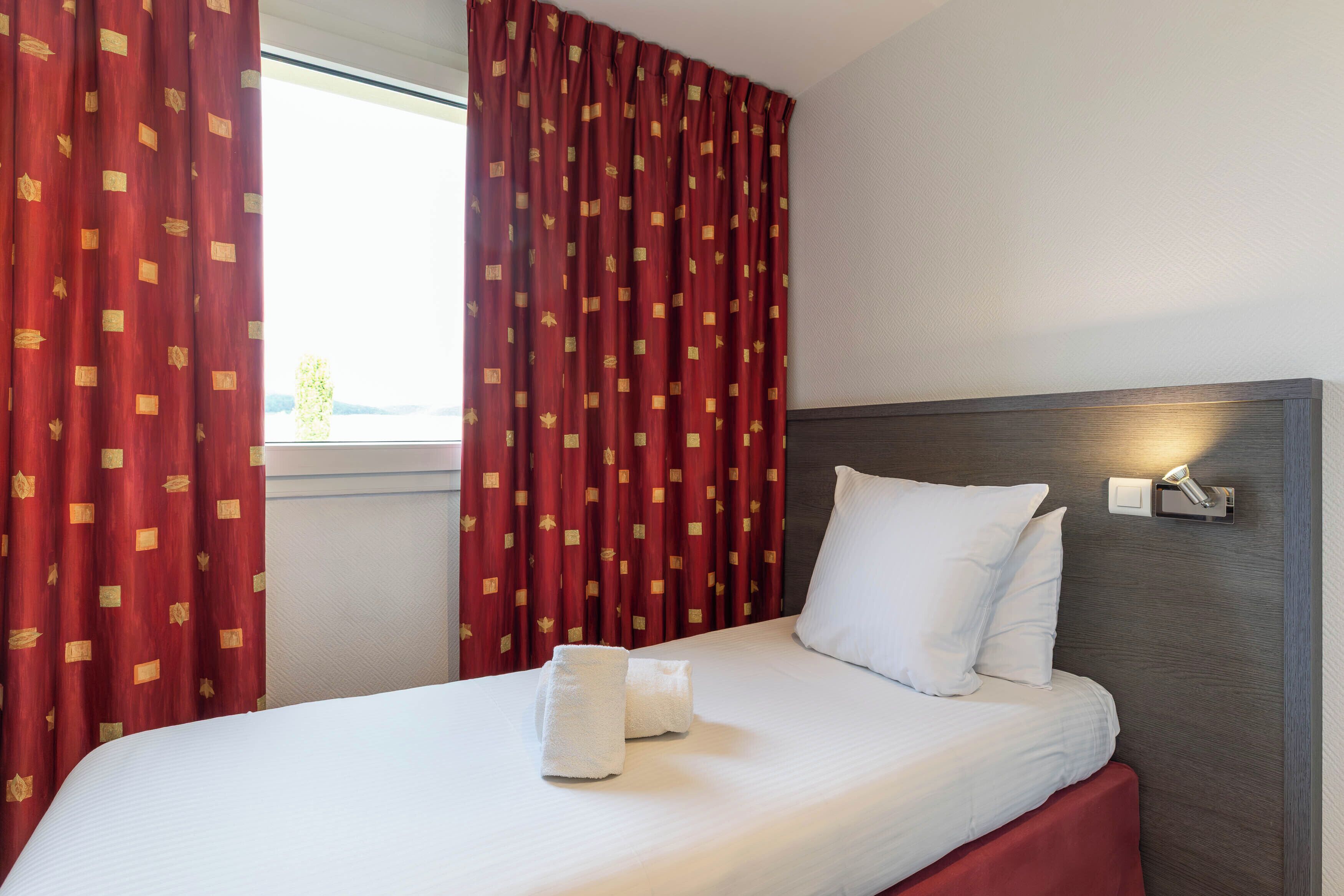 A hotel room with a bed, white pillows, red curtains with a pattern, a bedside lamp, and a window.