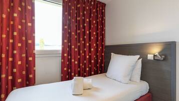 A hotel room with a bed, white pillows, red curtains with a pattern, a bedside lamp, and a window.