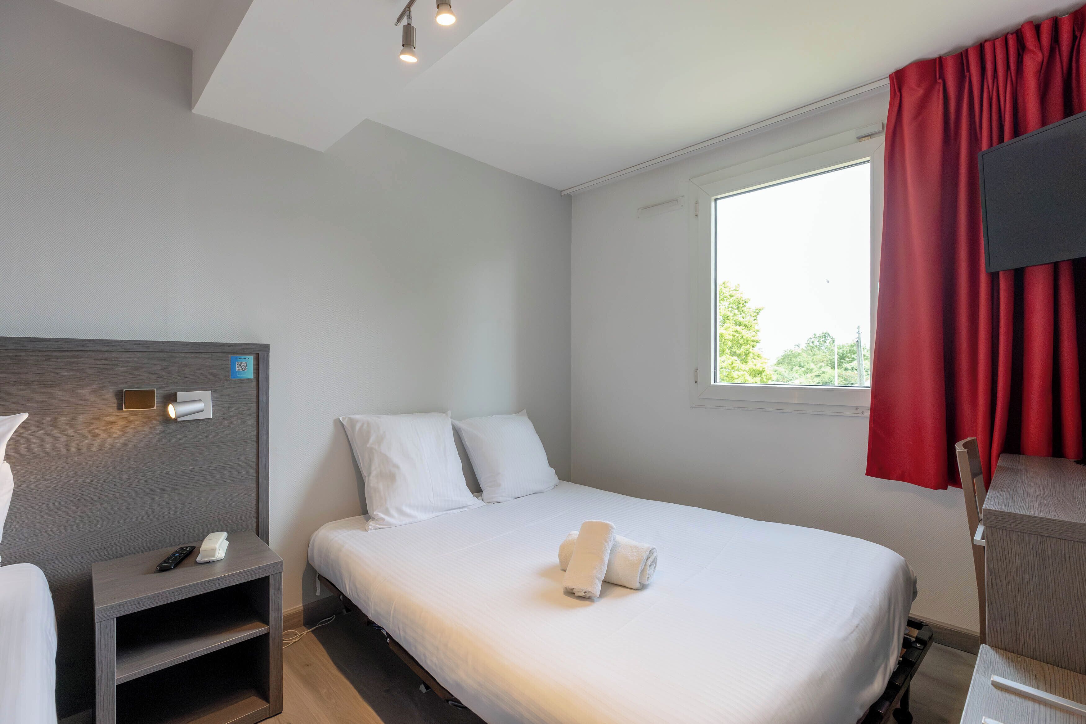 A hotel room with a bed, a nightstand with a phone, a TV mounted on the wall, and a window with red curtains.