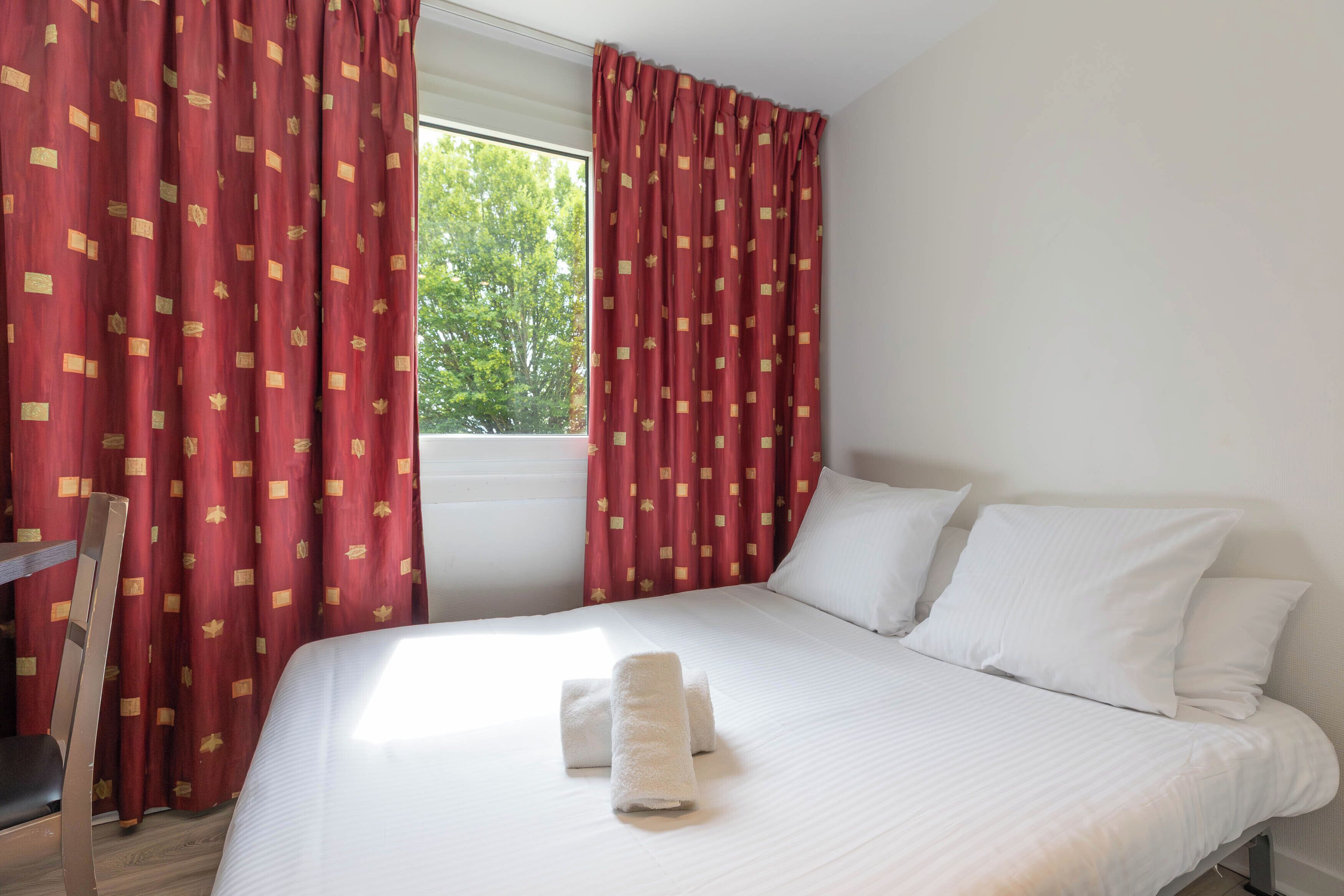 Quarto de hotel com cama, almofadas brancas e janela com cortinas vermelhas.