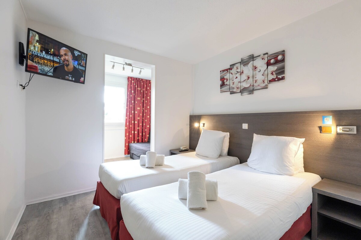 Une chambre d’hôtel avec deux lits, une télévision, une fenêtre avec des rideaux rouges et des œuvres d’art accrochées au mur.