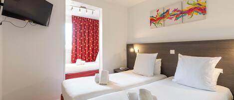 A hotel room with two beds, a TV mounted on the wall, a window with red curtains, and a colorful abstract painting on the wall.