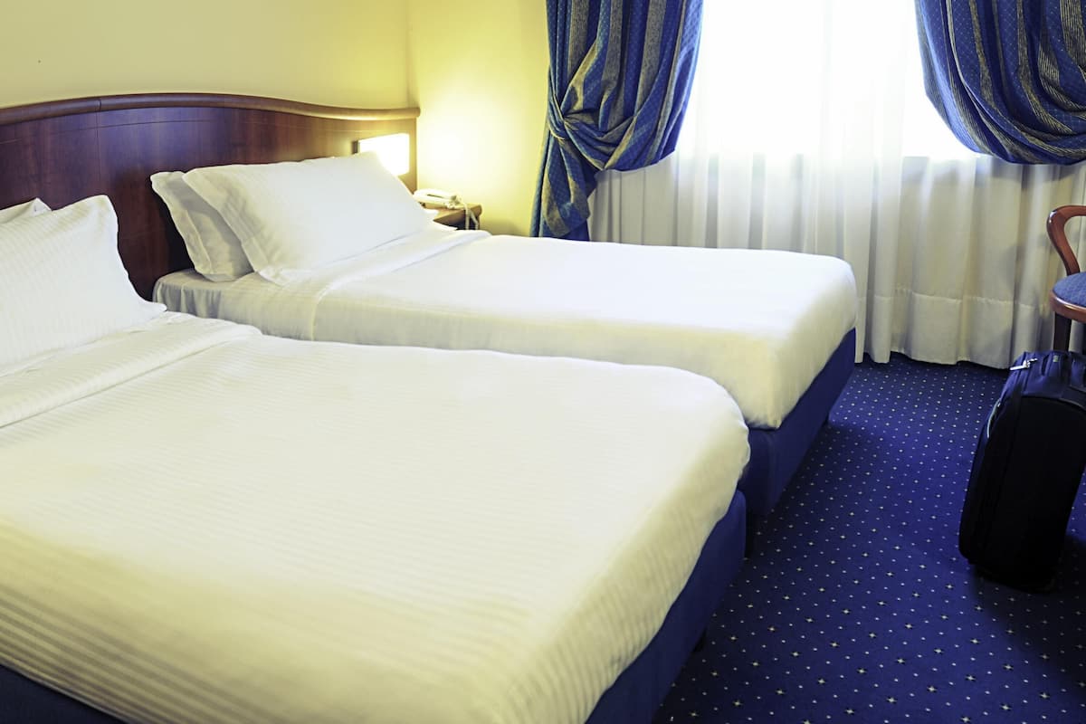 Two single beds with white linens, a wooden headboard, a nightstand, a suitcase, and a window with blue curtains.