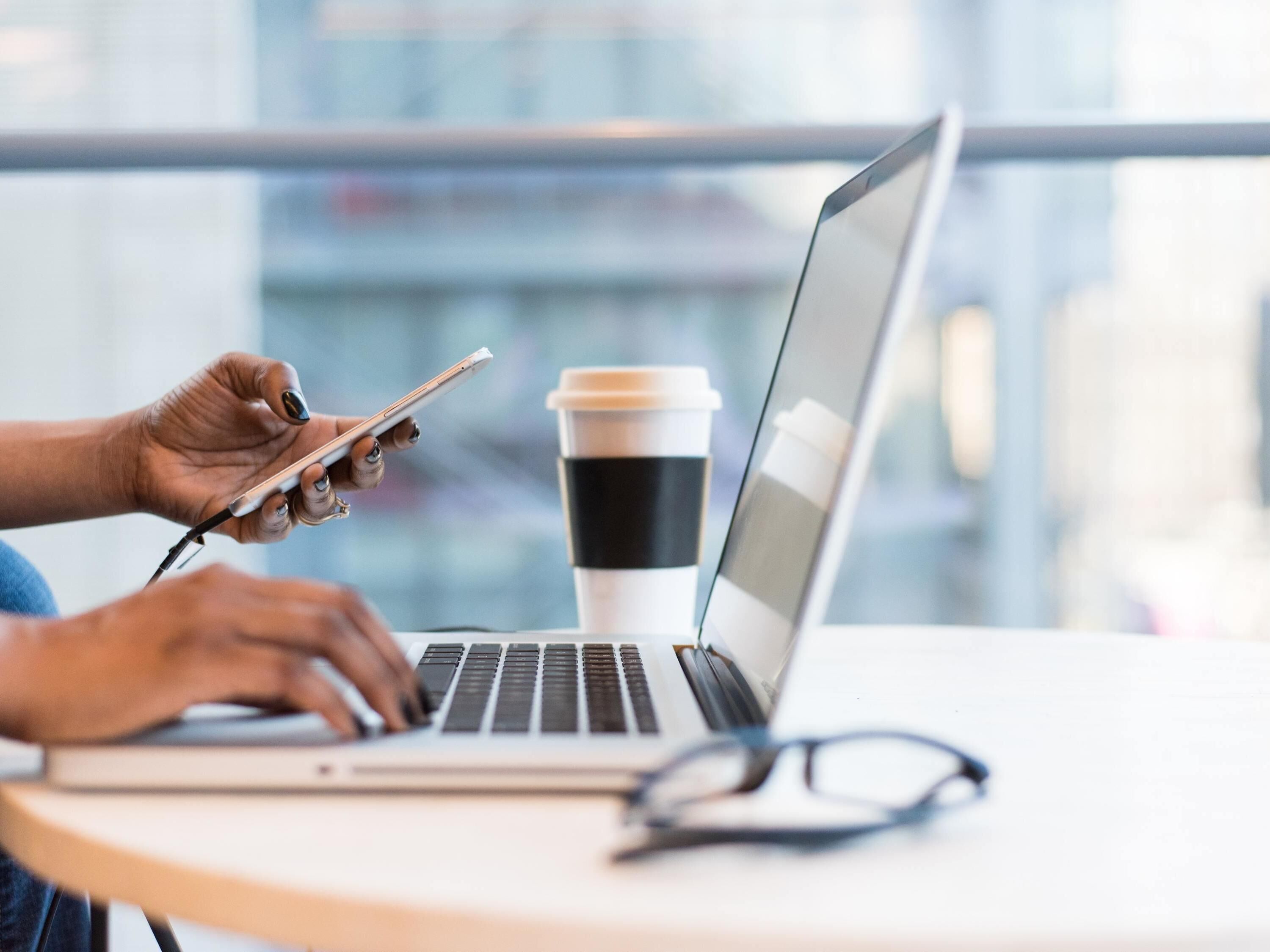 A person using a laptop with a smartphone and a coffee cup on a table.