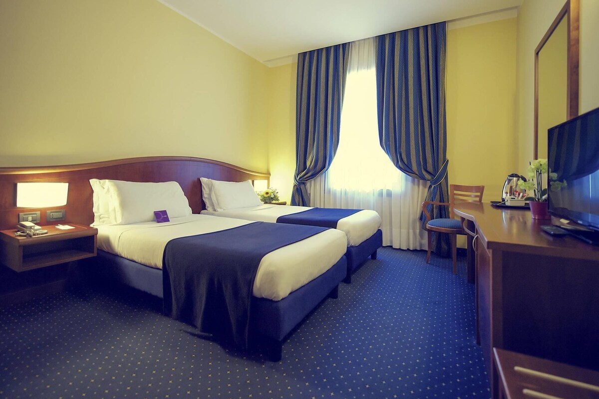 A hotel room with two beds, a desk with a chair, a television, and a window with curtains.