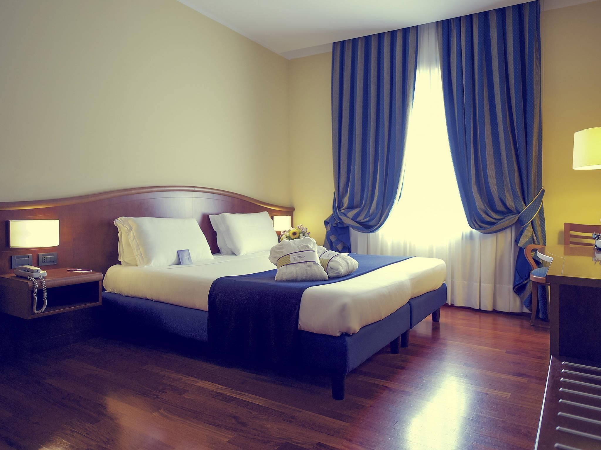 Ein Hotelzimmer mit einem hölzernen Bett, einem Nachttisch mit Lampe, einem Schreibtisch mit Stuhl und einem großen Fenster mit blauen Vorhängen.
