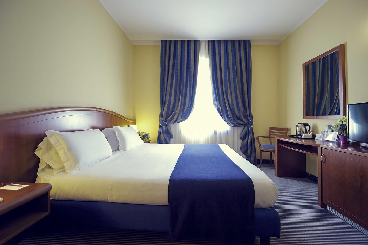A hotel room with a bed, a desk, a chair, a television, and a large window with curtains.