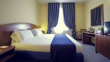 Camera d'albergo con un letto, una scrivania, una sedia, una televisione e un'ampia finestra con tende.