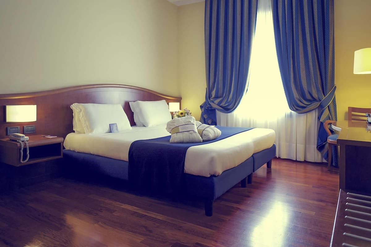 A hotel room with a wooden bed, a nightstand with a phone, a desk with a lamp, and a large window with curtains.