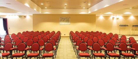 A conference room with rows of red chairs, a projector on the ceiling, and a carpeted floor.