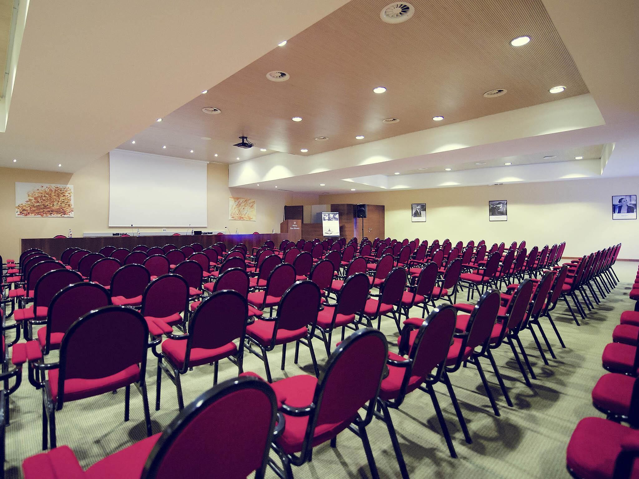 Konferenssisali, jossa on riveissä punaisia tuoleja, puhujapönttö, näyttö ja kattoon upotettu valaistus.