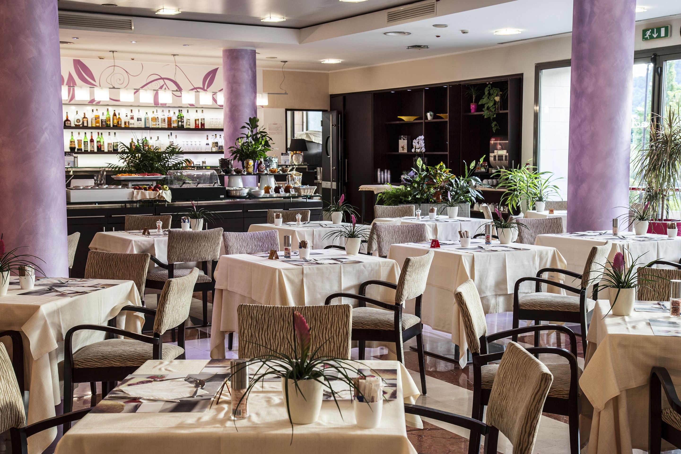 A restaurant with multiple tables and chairs, a bar area with bottles, and potted plants.