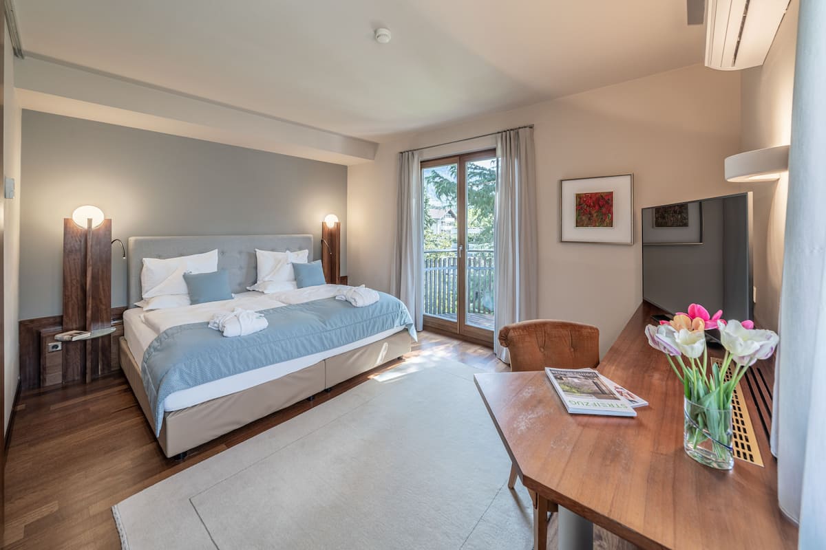 Habitación de hotel moderna con una cama grande, mesitas de noche, un escritorio de madera, una silla y una ventana con vistas a zonas verdes.