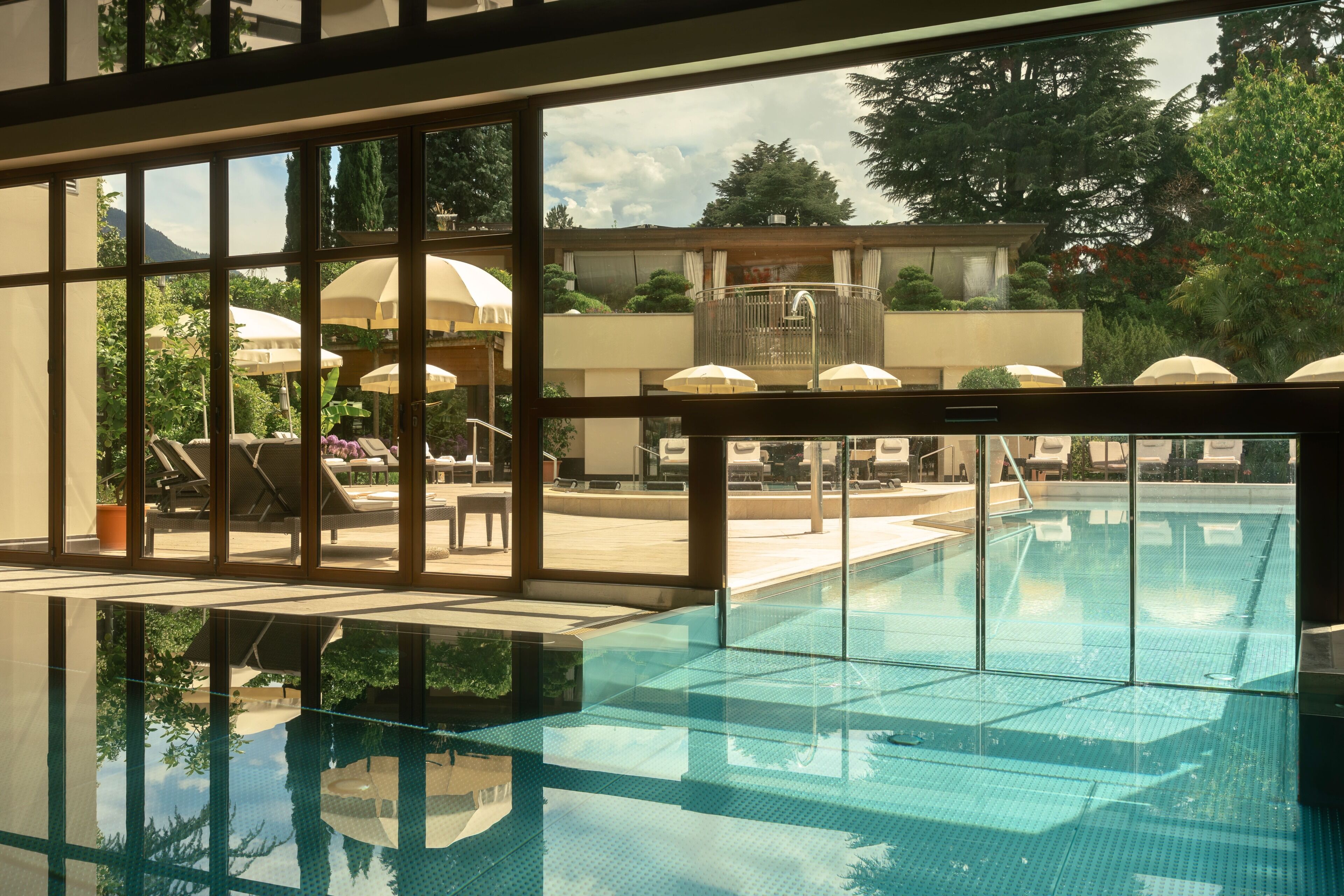 Ein Schwimmbecken mit klarem Wasser, umgeben von Liegestühlen und Sonnenschirmen, mit Blick auf Bäume und ein Gebäude im Hintergrund.