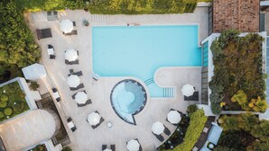 An aerial view of a swimming pool area with lounge chairs and umbrellas, surrounded by greenery and a tiled deck.