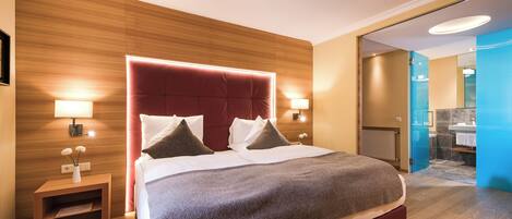 Une chambre d’hôtel moderne avec un grand lit, des murs recouverts de panneaux en bois, une table de chevet avec une lampe et une salle de bain visible à travers une porte ouverte.