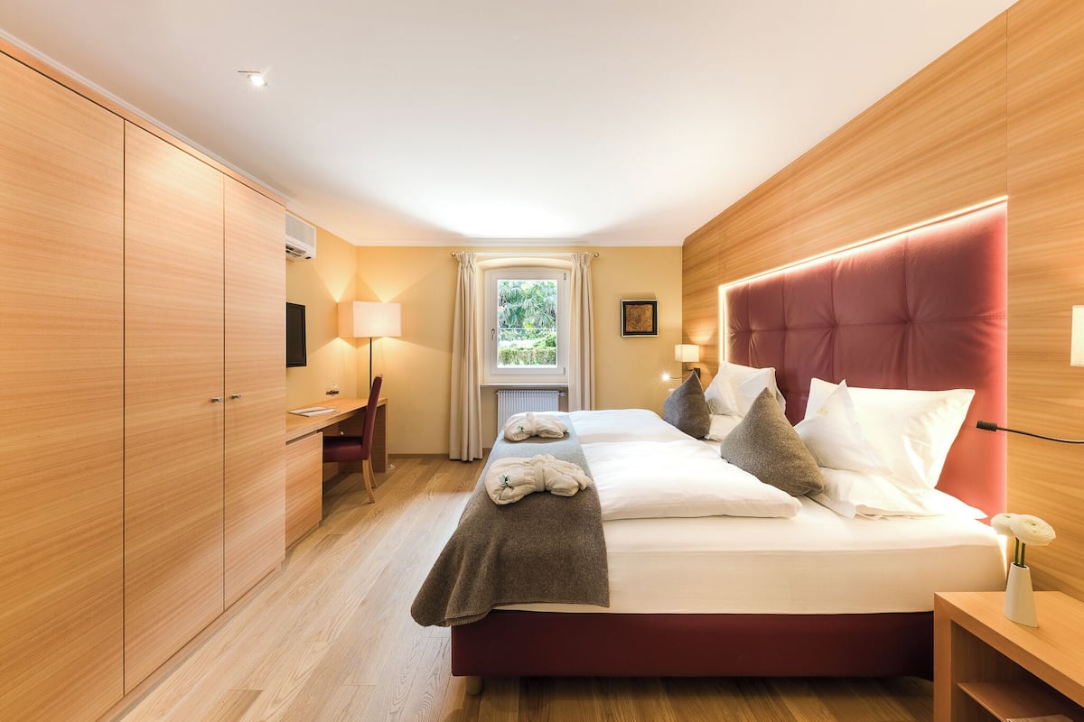 Habitación de hotel con cama, mesitas de noche, escritorio, silla, ventana con cortinas y vista a zonas verdes.
