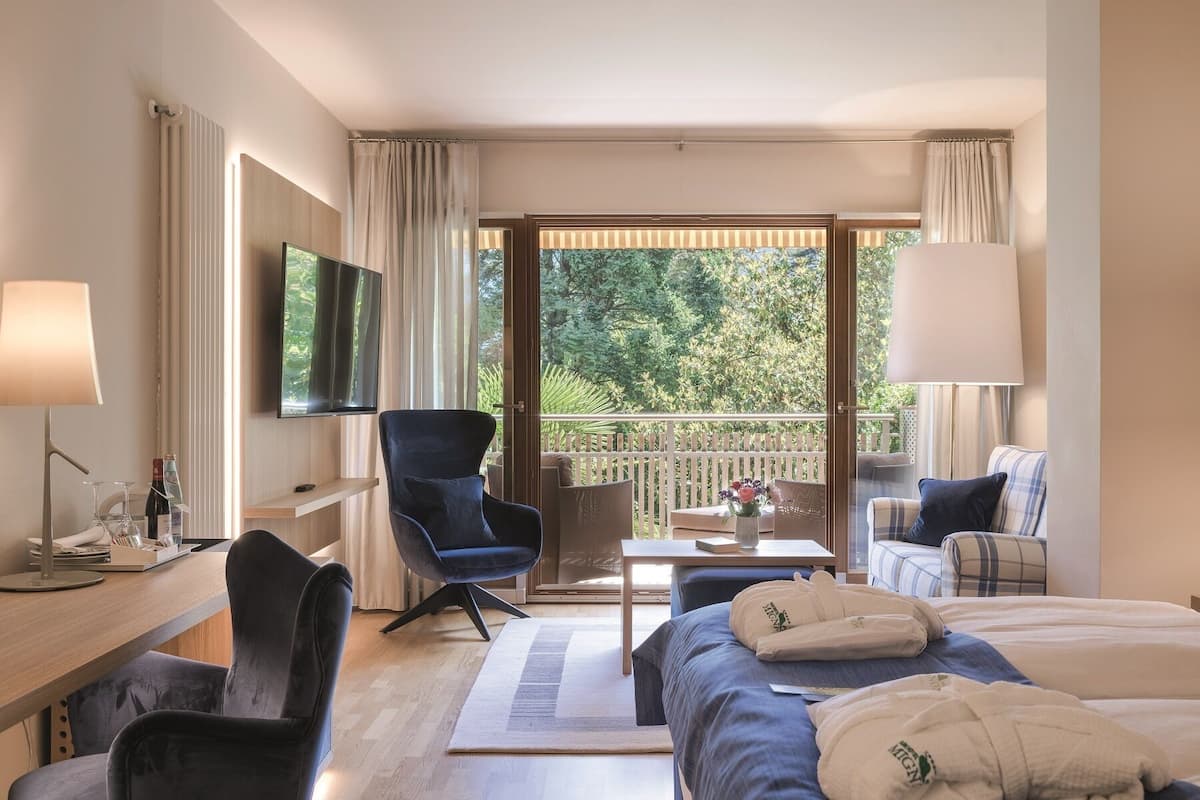 Habitación de hotel con cama, escritorio, silla, televisión y balcón con vistas a árboles.