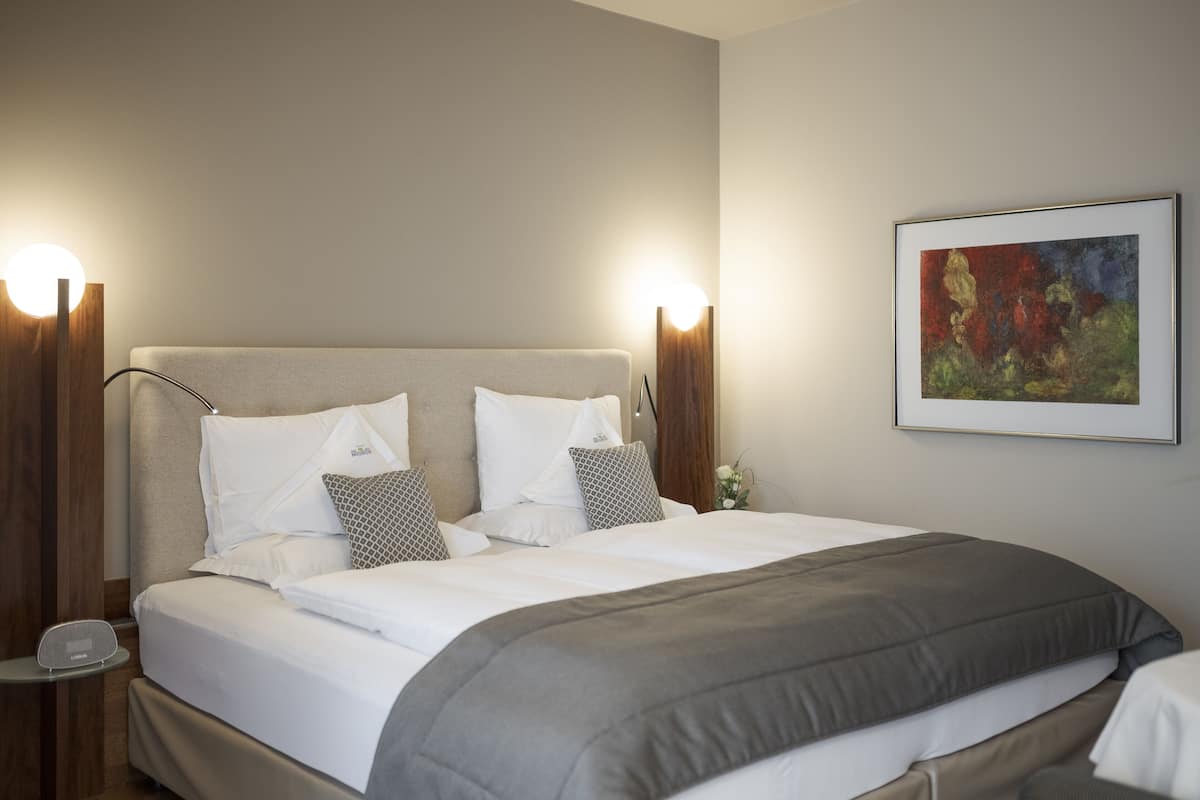 Una cama bien hecha con sábanas blancas y una manta gris, flanqueada por dos mesitas de noche con lámparas, en una habitación con un cuadro en la pared.