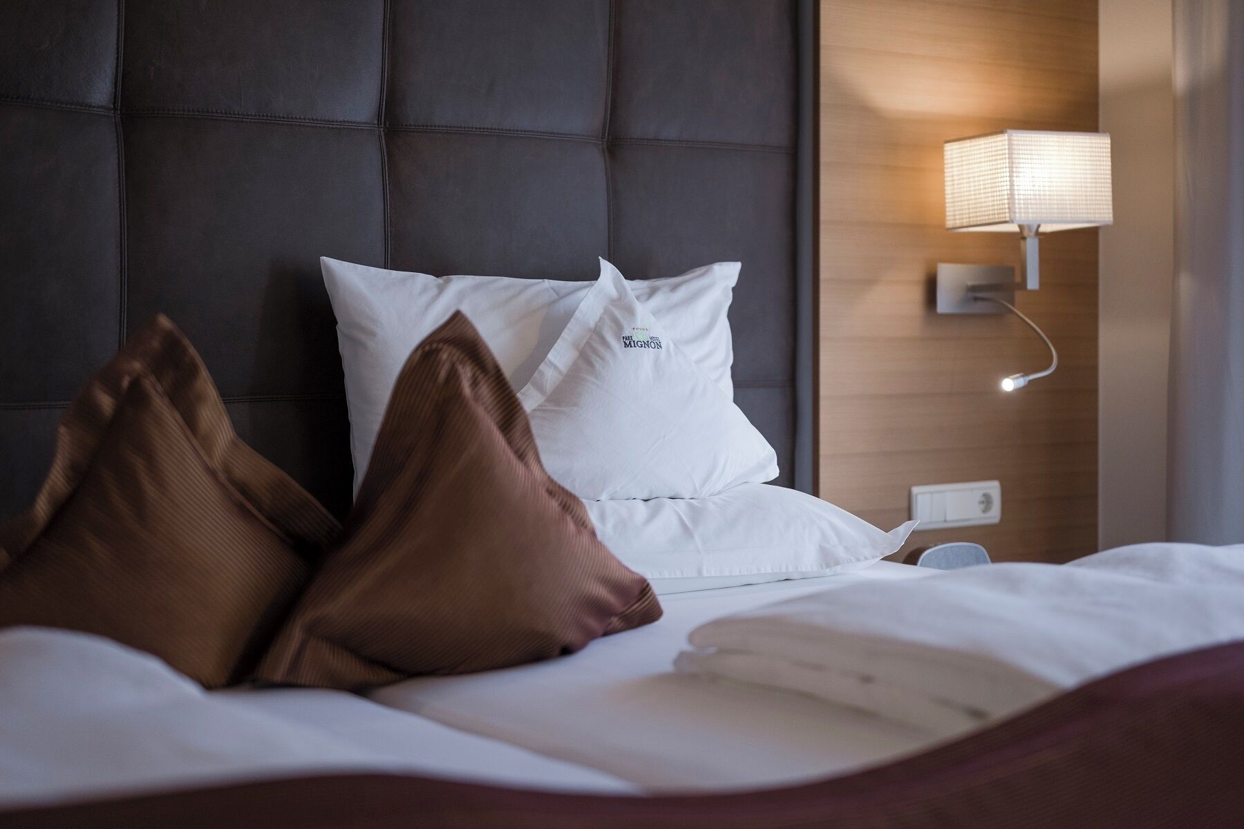 Ein Bett mit weißer Bettwäsche, braunen Kissen, einem dunklen Kopfteil und einer Wandleuchte.
