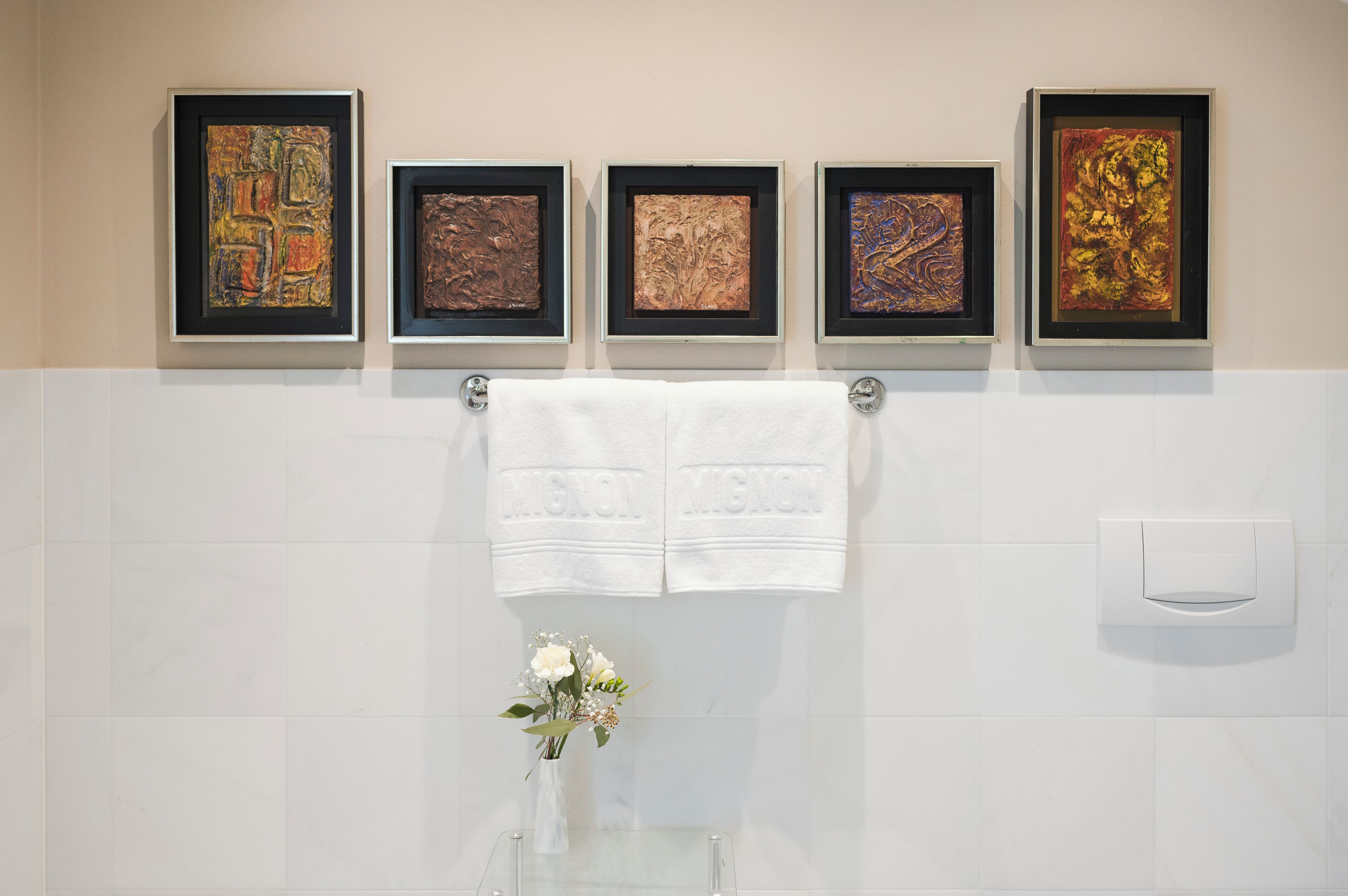 Ein Badezimmer mit fünf gerahmten Kunstwerken an der Wand, zwei weißen Handtüchern und einer Vase mit Blumen auf der Ablagefläche.