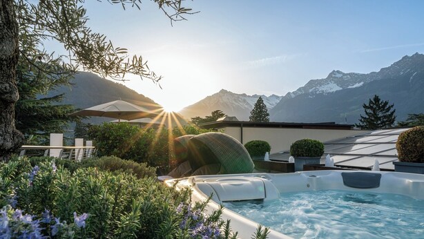A hot tub with a mountain view, surrounded by lush greenery and a white fence.