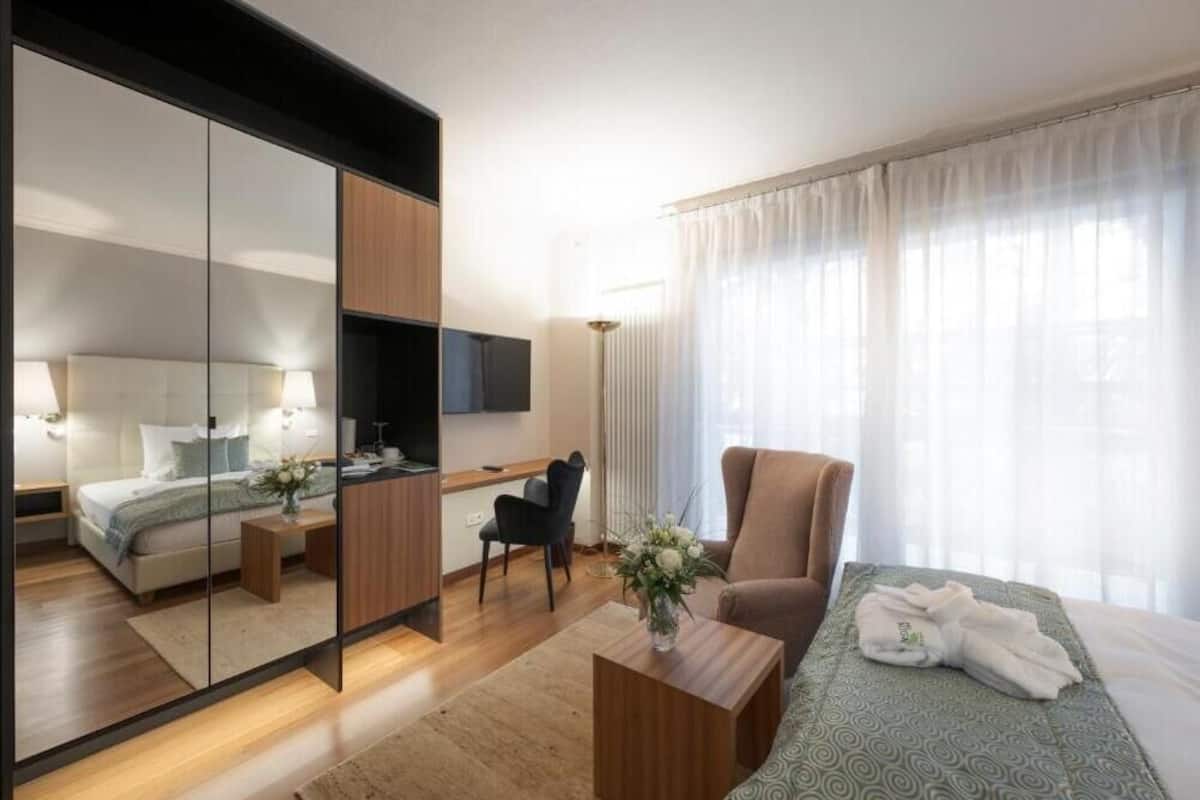 Una habitación de hotel moderna con un espejo grande, una cama con colcha verde estampada, una zona de estar con una silla y una mesita, y un escritorio con silla.