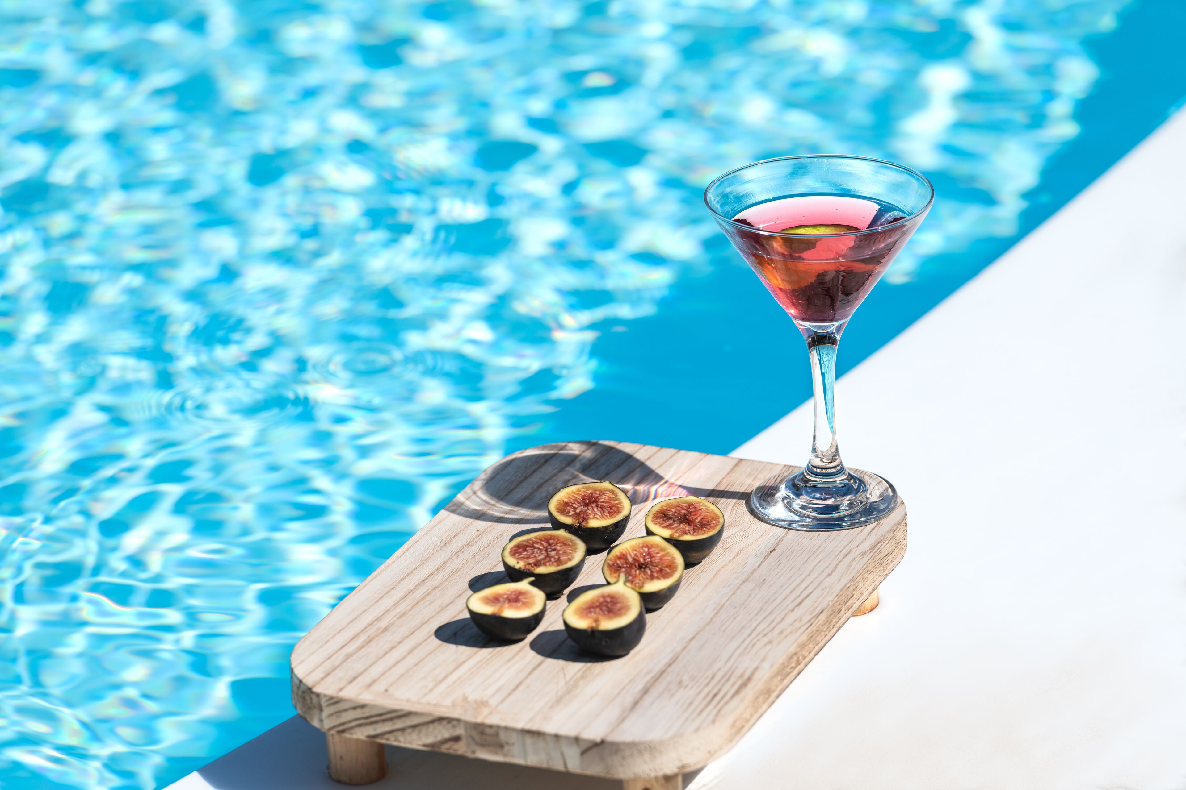 A wooden table with a glass of pink drink and sliced figs by a pool.
