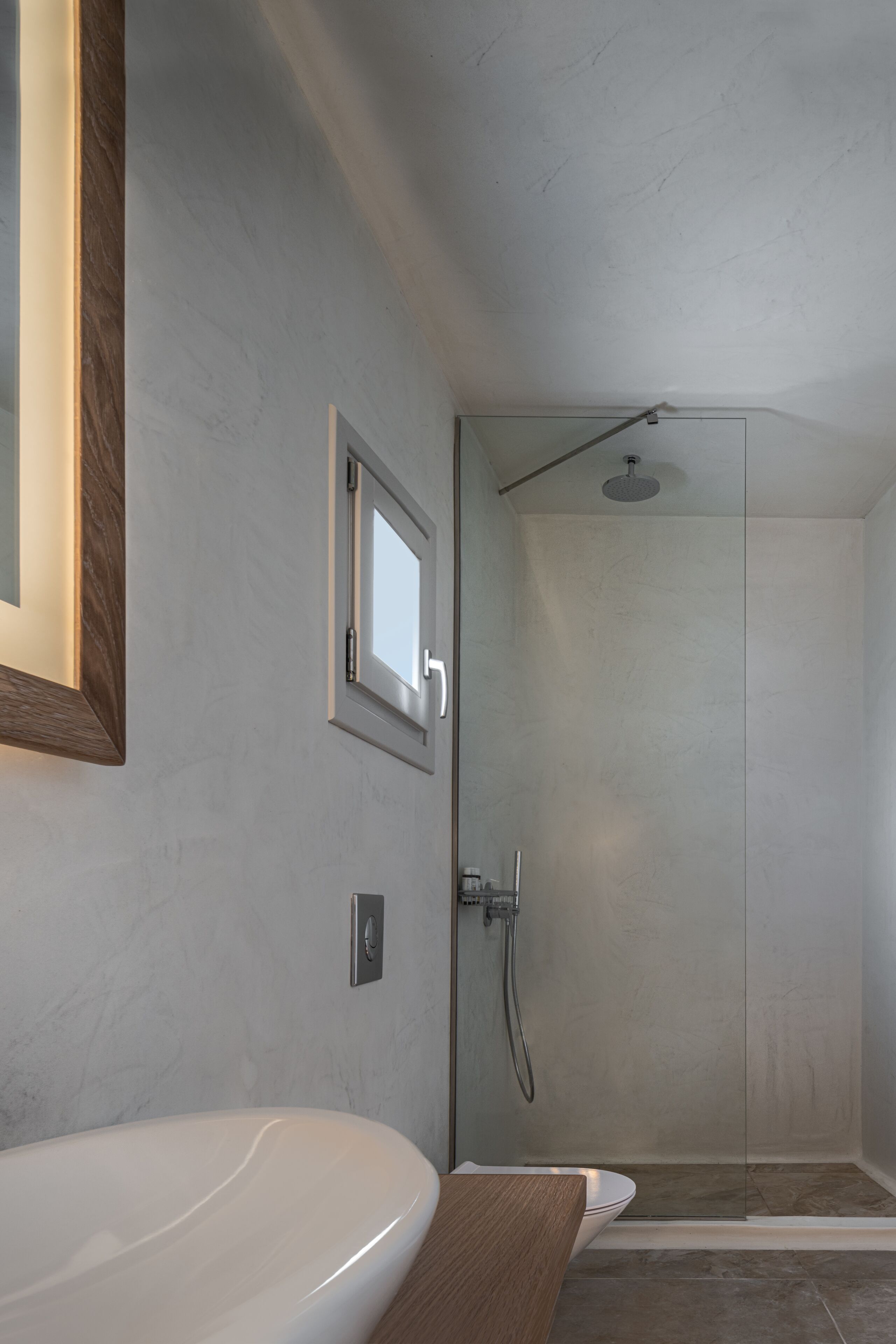 A modern bathroom with a white sink, a wooden countertop, a glass shower enclosure, and a small window.