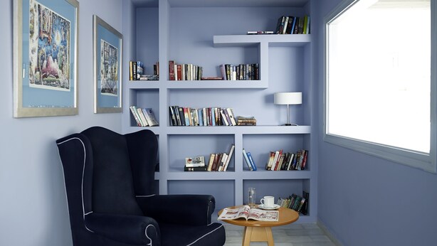 A room with a dark blue armchair, a wooden coffee table with a magazine, and a bookshelf filled with books. - Santorini Kastelli Resort (Santorini)