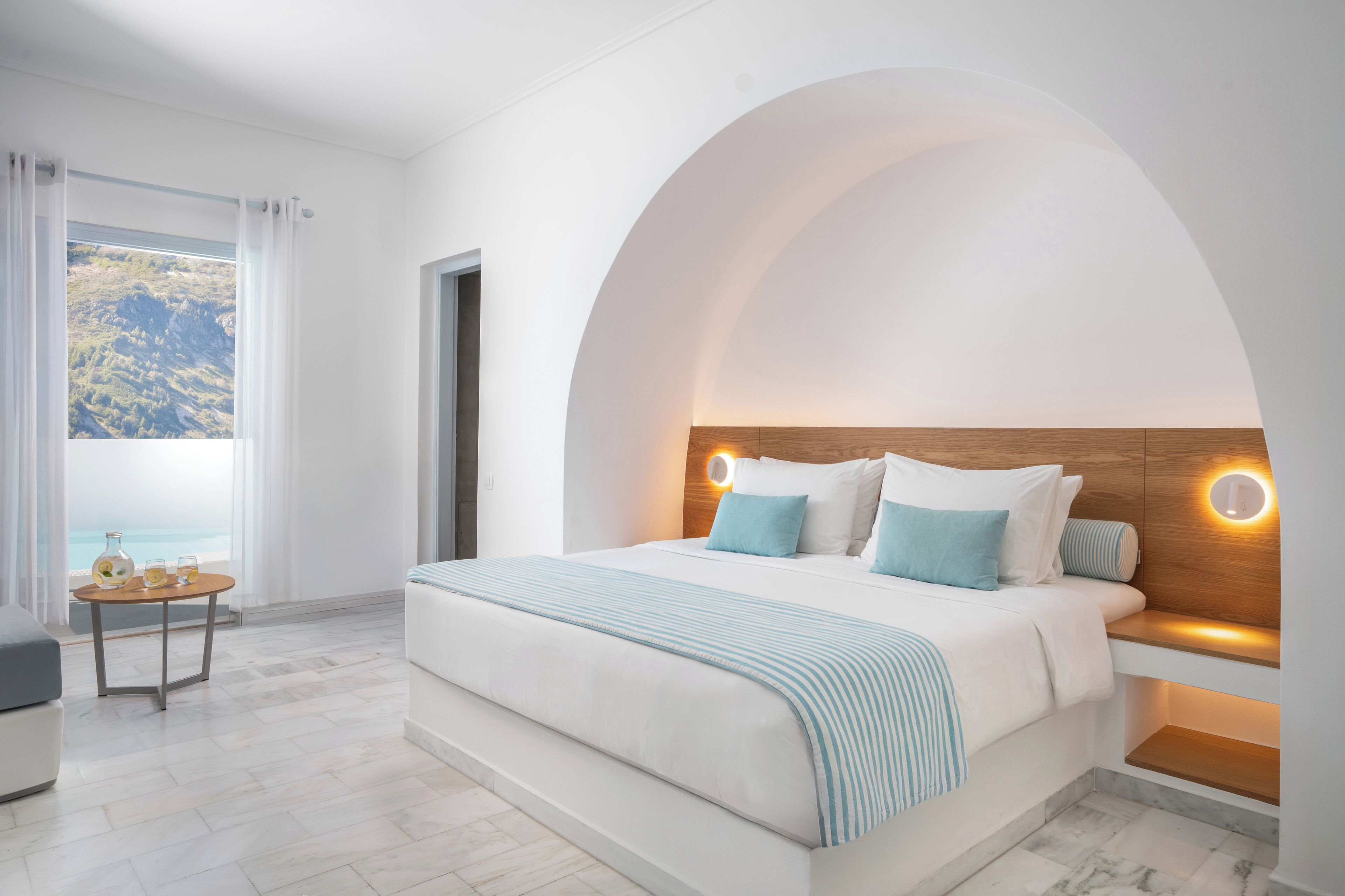 Een moderne slaapkamer met een groot bed, een nachtkastje met een lamp, uitzicht op zee door een raam, en een klein tafeltje met drankjes.