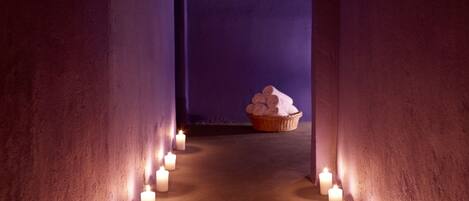 A hallway with a row of lit candles on the floor, leading to a room with a basket of towels.