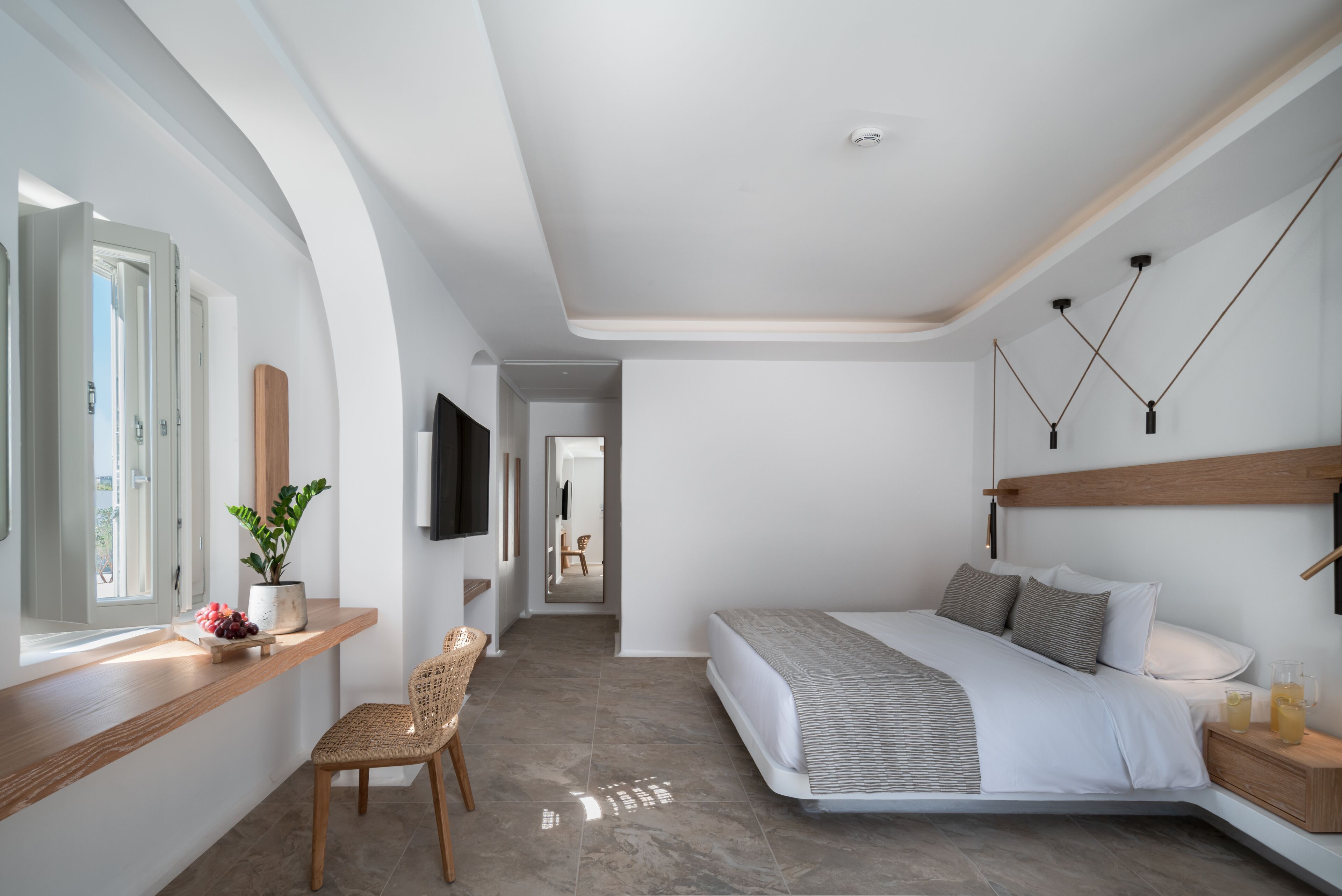 A modern bedroom with a curved bed, a wooden bedside table, a chair, a TV mounted on the wall, and a window with a view of the outdoors.