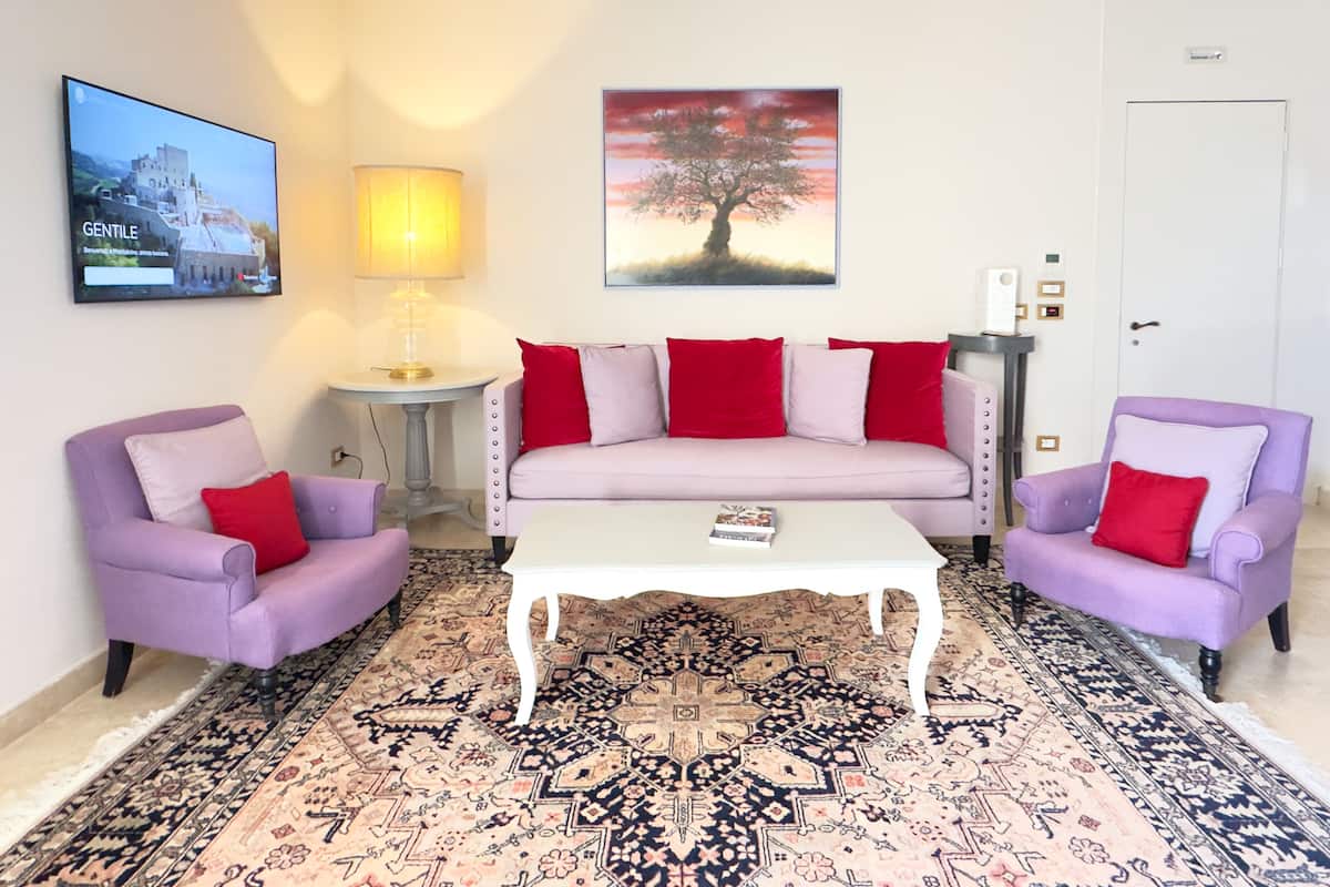 A living room with a sofa, two armchairs, a coffee table, a TV, a painting, and a patterned rug.