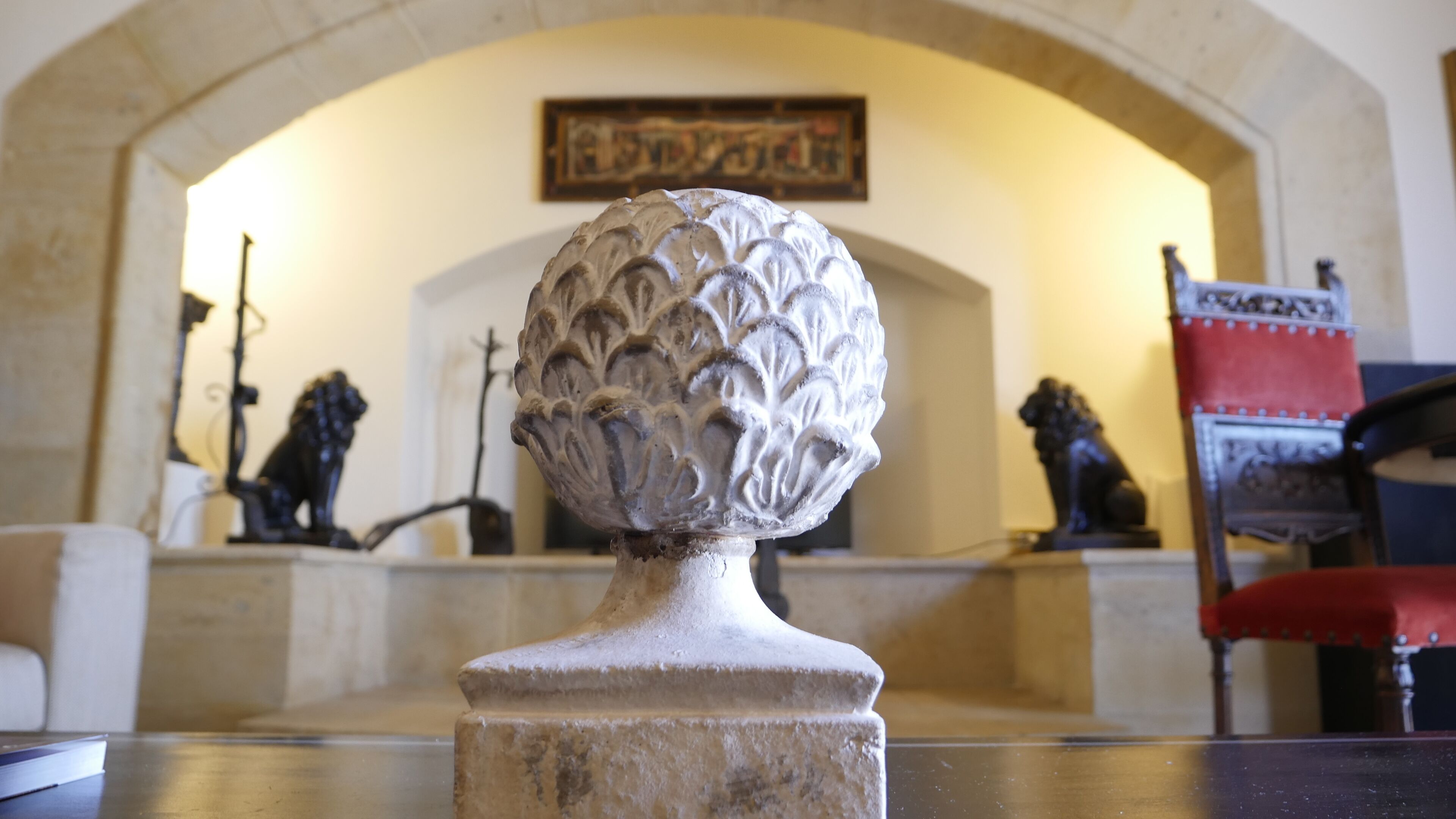 A stone bust with intricate carvings, placed on a pedestal in a room with a high arched ceiling and a framed artwork on the wall.
