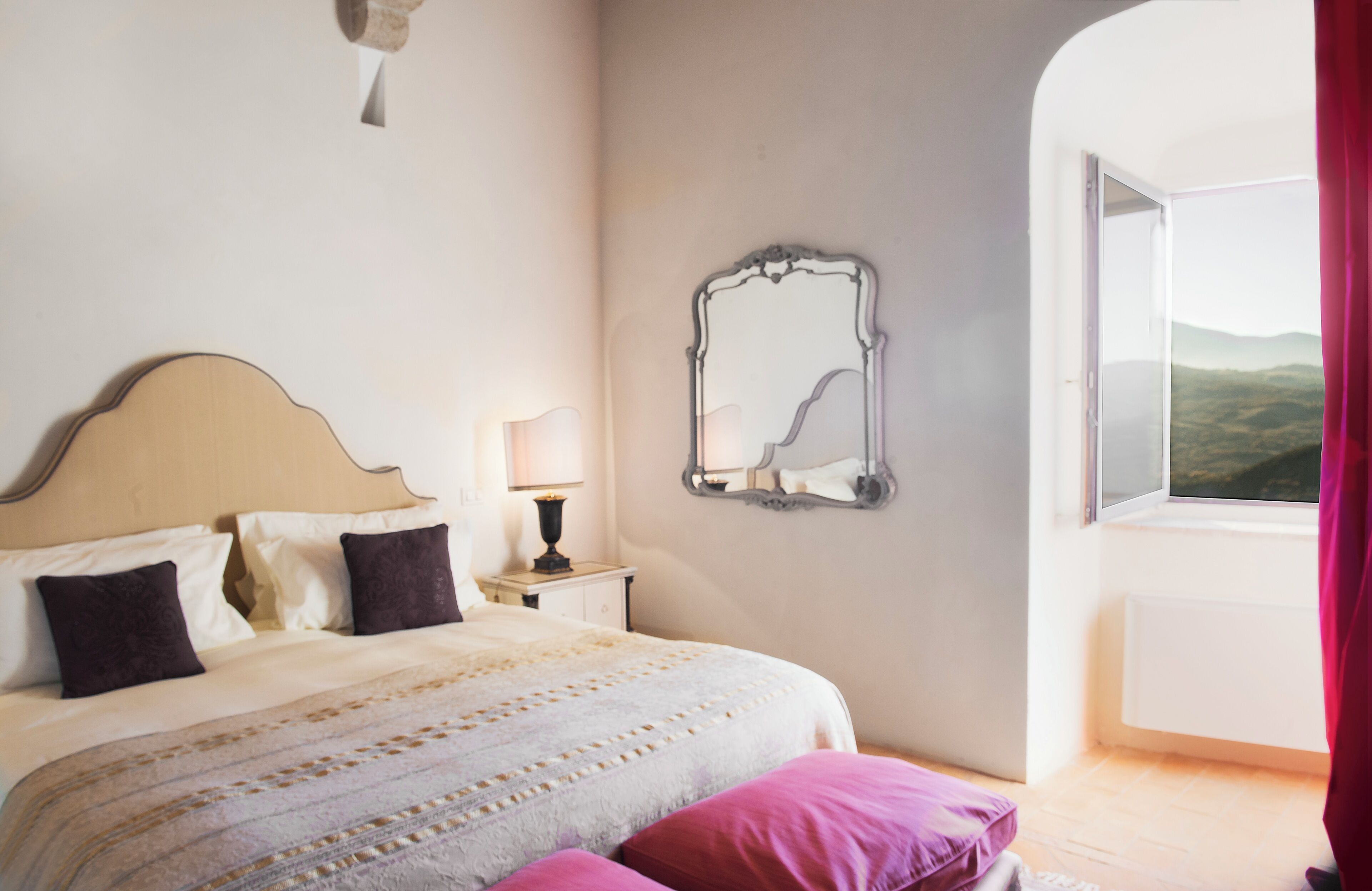 Een slaapkamer met een bed, kussens, een nachtkastje, een lamp, een spiegel en een raam met uitzicht.