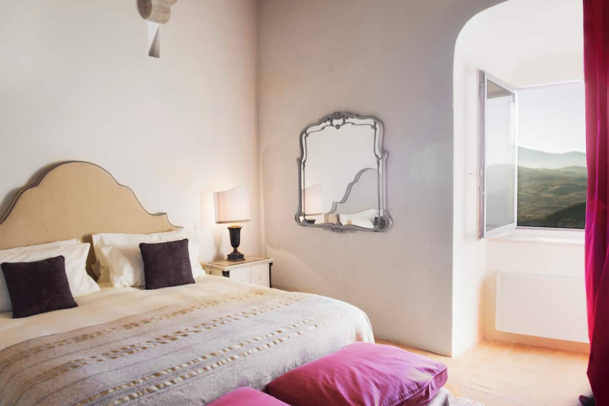 A bedroom with a bed, pillows, a nightstand, a lamp, a mirror, and a window with a view.