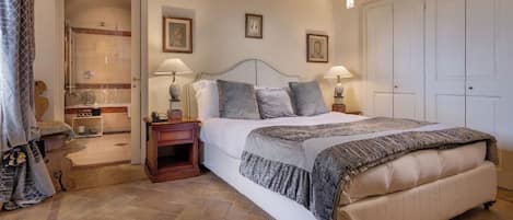 A bedroom with a large bed, two bedside tables with lamps, a wooden nightstand, a wardrobe, and a bathroom with a shower and toilet visible.