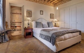 A bedroom with a large bed, two bedside tables with lamps, a wooden nightstand, a wardrobe, and a bathroom with a shower and toilet visible. - Castello di Velona Resort Thermal SPA & Winery (Montalcino)