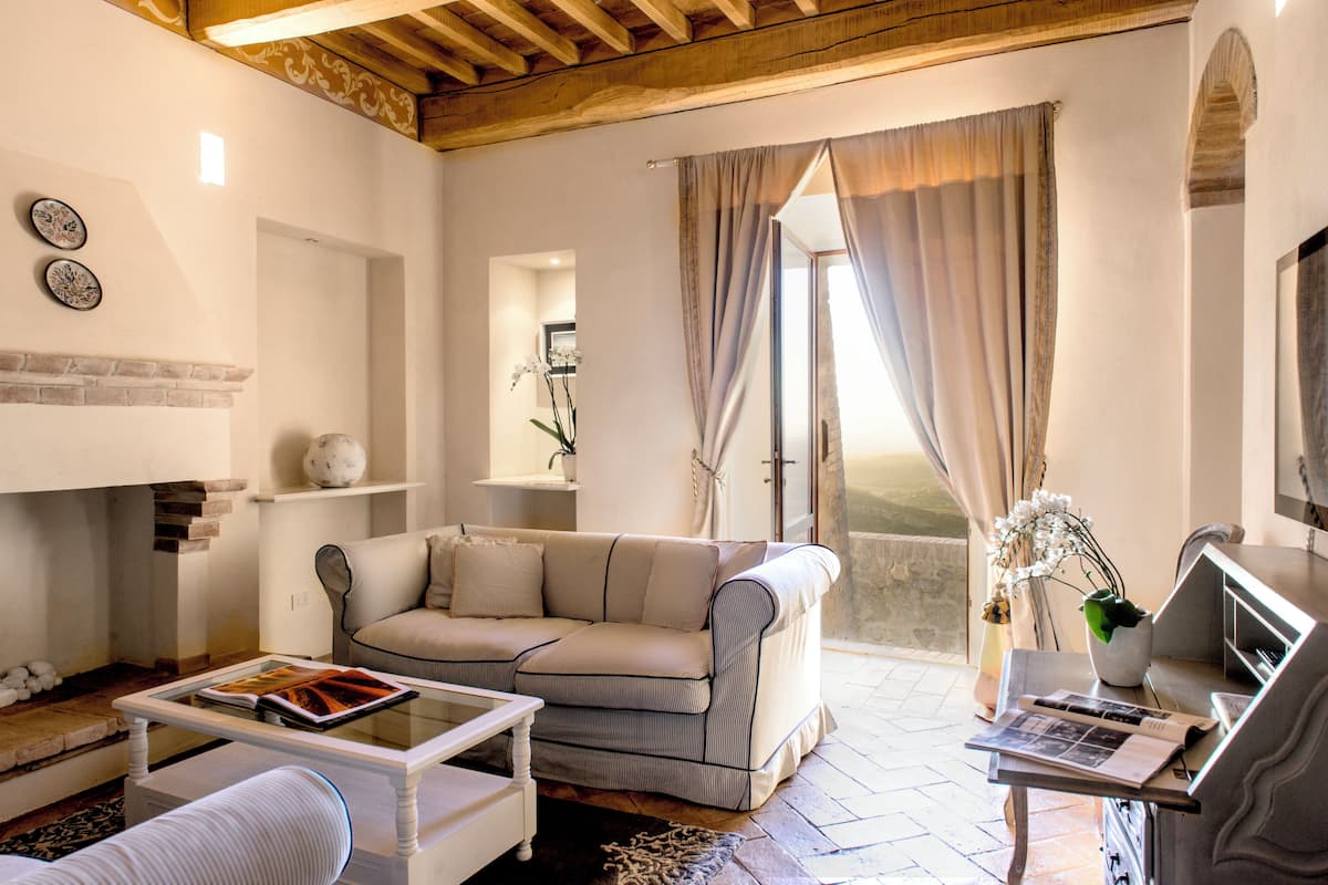 A living room with a sofa, coffee table, and a fireplace. There is a window with curtains and a view of the outside.