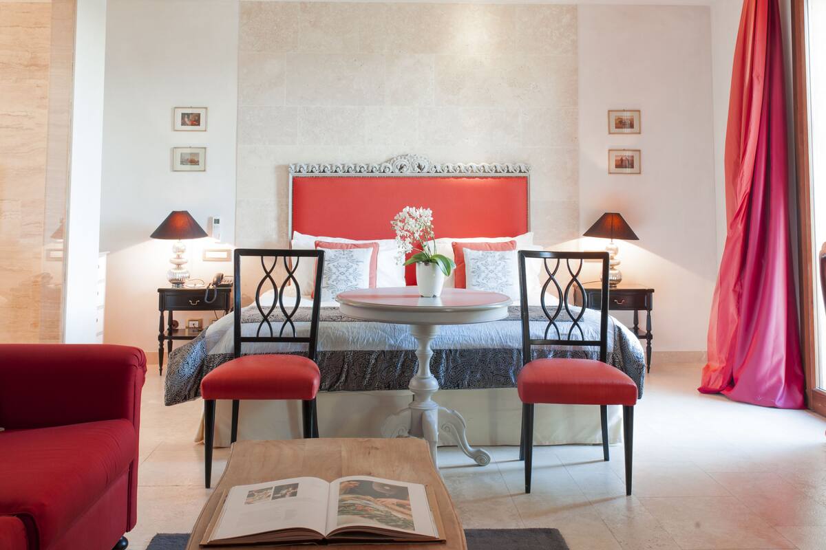 A hotel room with a bed, a round table with chairs, a red sofa, and two bedside tables with lamps.