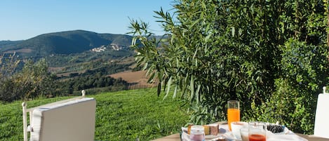 A table set outdoors with a view of rolling hills, a chair, and a breakfast spread including croissants, cheese, and juice.