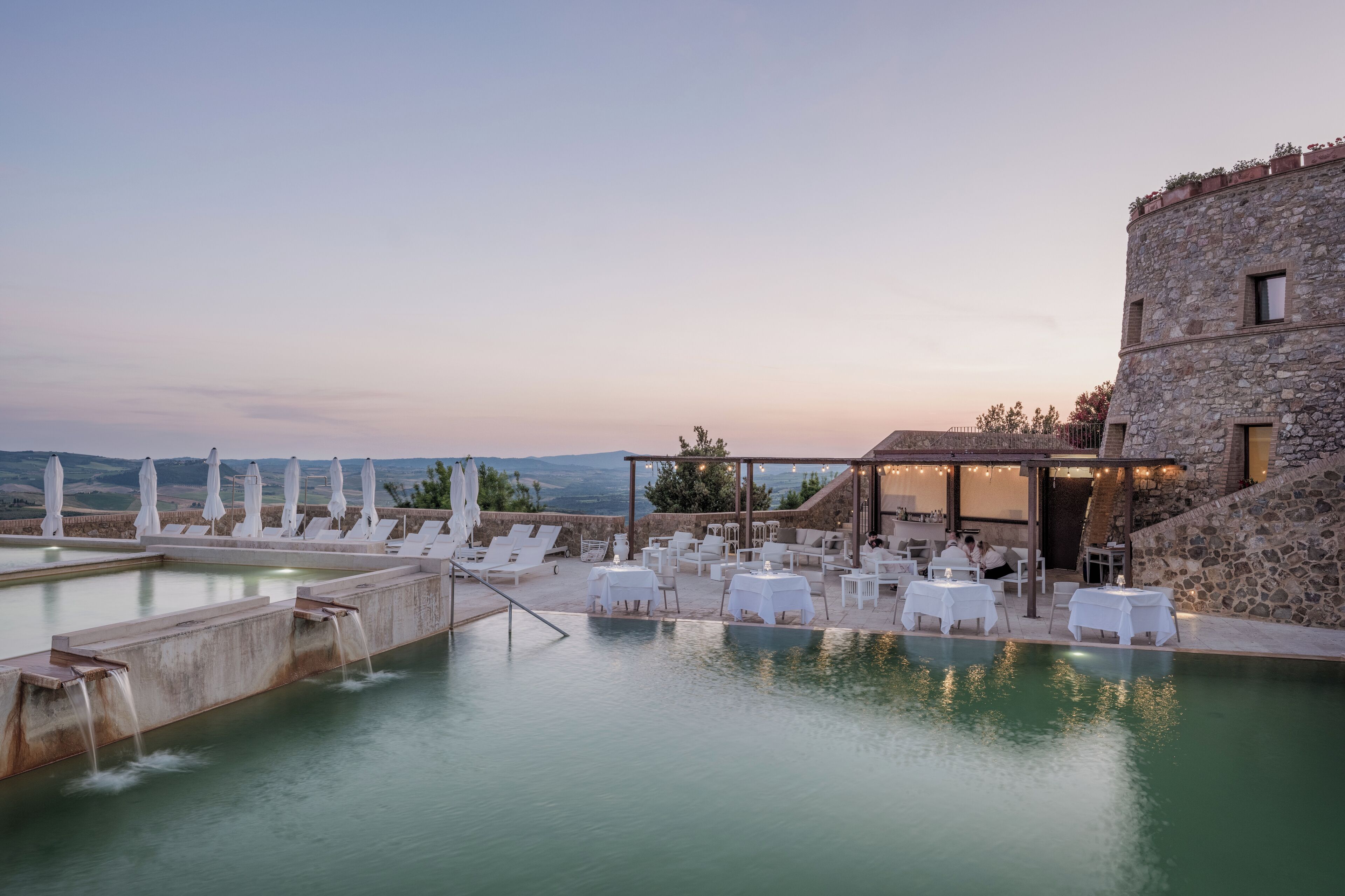 A rooftop pool with lounge chairs and tables, surrounded by stone walls and a view of the landscape.